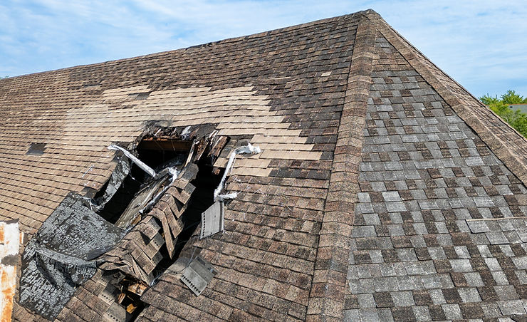 Damaged roof with missing and broken shingles creating a large hole. The shingles are brown and gray. Bright blue sky in the background.